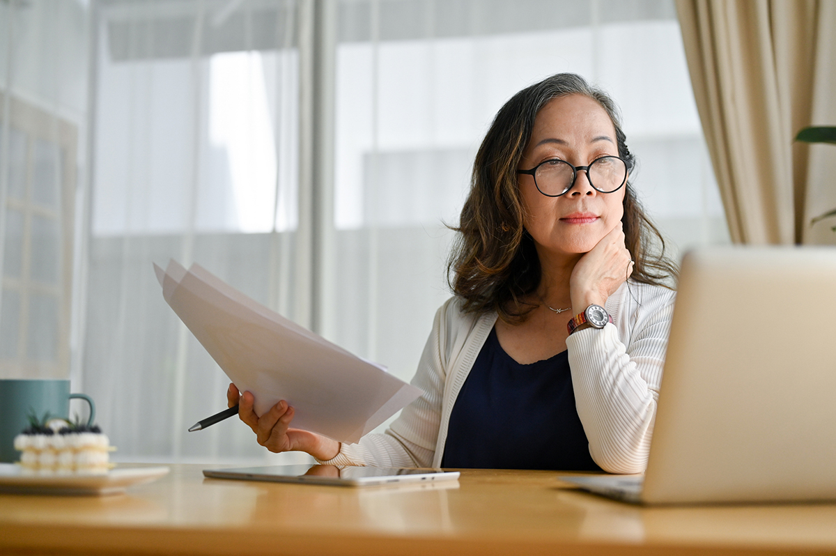 Asian-American woman reviewing mortgage estimates on laptop, papers and pen in hand, sitting at table with natural light from picture window in background.