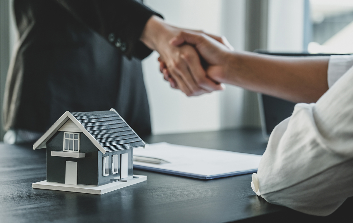 Real estate agent shaking hands with a client over a desk with a model house and paperwork.