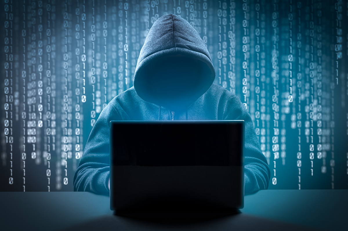 A hooded figure at a laptop, symbolizing the threat of cybercrime and digital scams in real estate transactions.